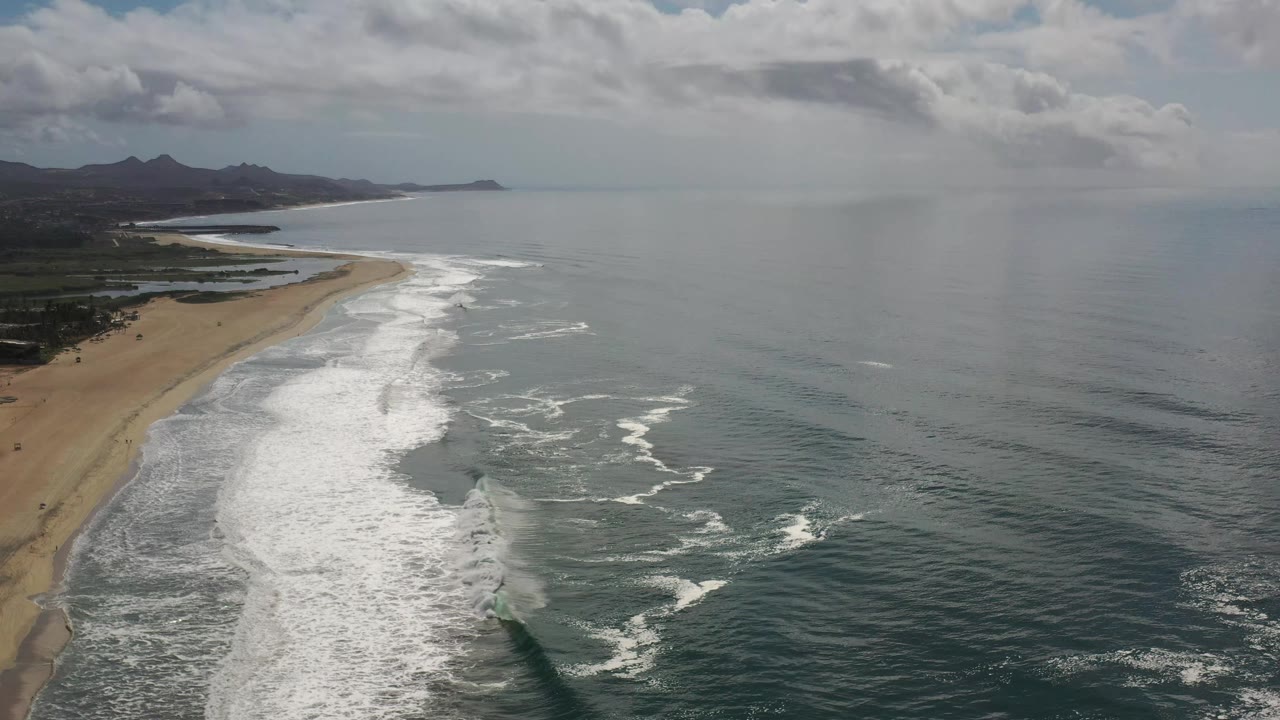 Aerial view in Los Cabos, Mexico showing more open water to the right, a curved sandy beach to the left, layered white surf, low hills on the horizon, and no visible people.