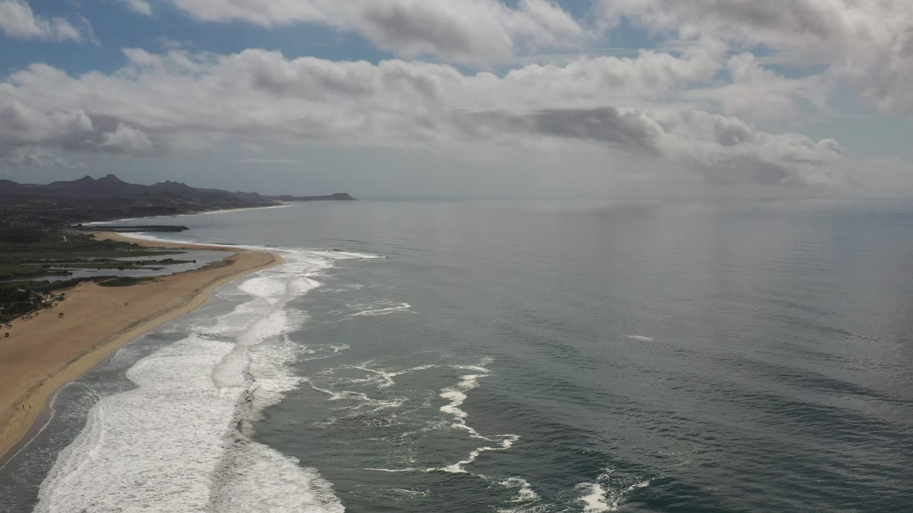 Aerial view over the Los Cabos coastline with tan beach along the left edge, white surf bands, open gray-green ocean, distant hills, layered clouds, and no visible people.