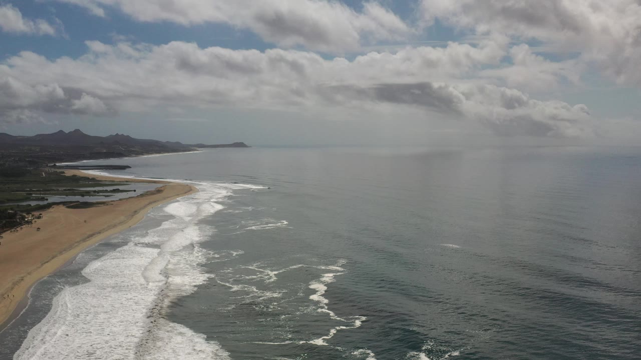 Drone-like view of beach surf, calm sea, distant headland, and layered clouds.