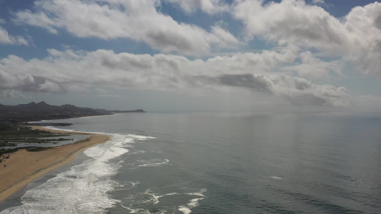 Aerial coastline with sandy beach, surf, ocean, distant hills, and clouded sky.