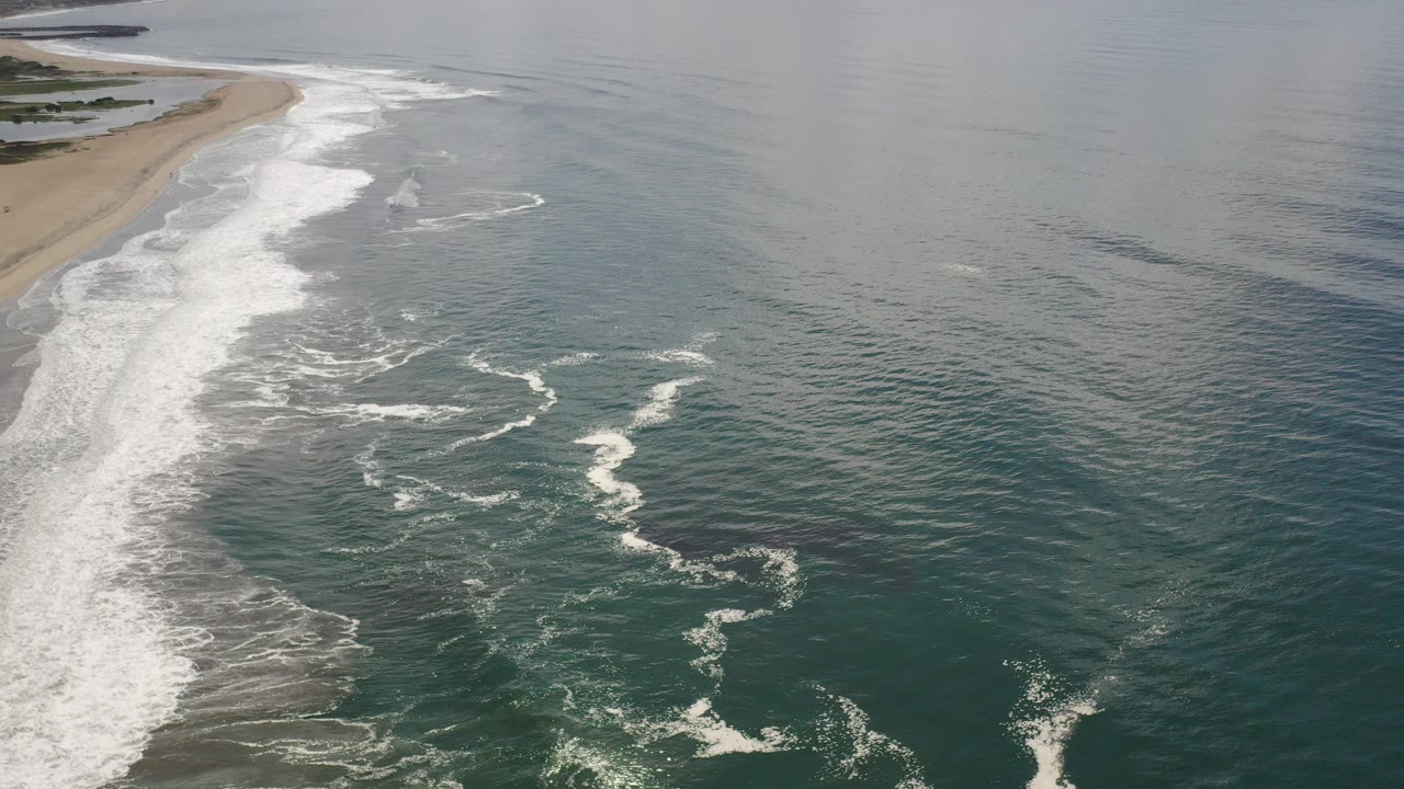 Aerial view over a Los Cabos beach with sand along the left edge, white surf curling through the center, green-blue ocean to the right, and no visible people.