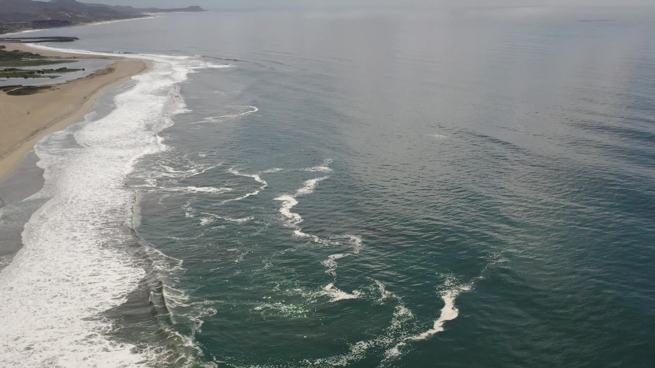 Los Cabos aerial frame with foamy wave bands advancing toward the sand, open ocean to the right, distant headland, and no visible people.