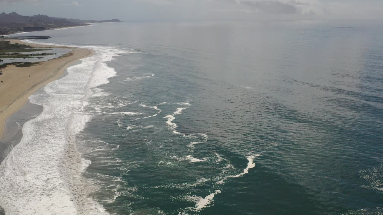 Opening aerial frame over a Los Cabos beach, with white surf curling along sand at left, open ocean to the right, distant hills on the horizon, and no visible people.