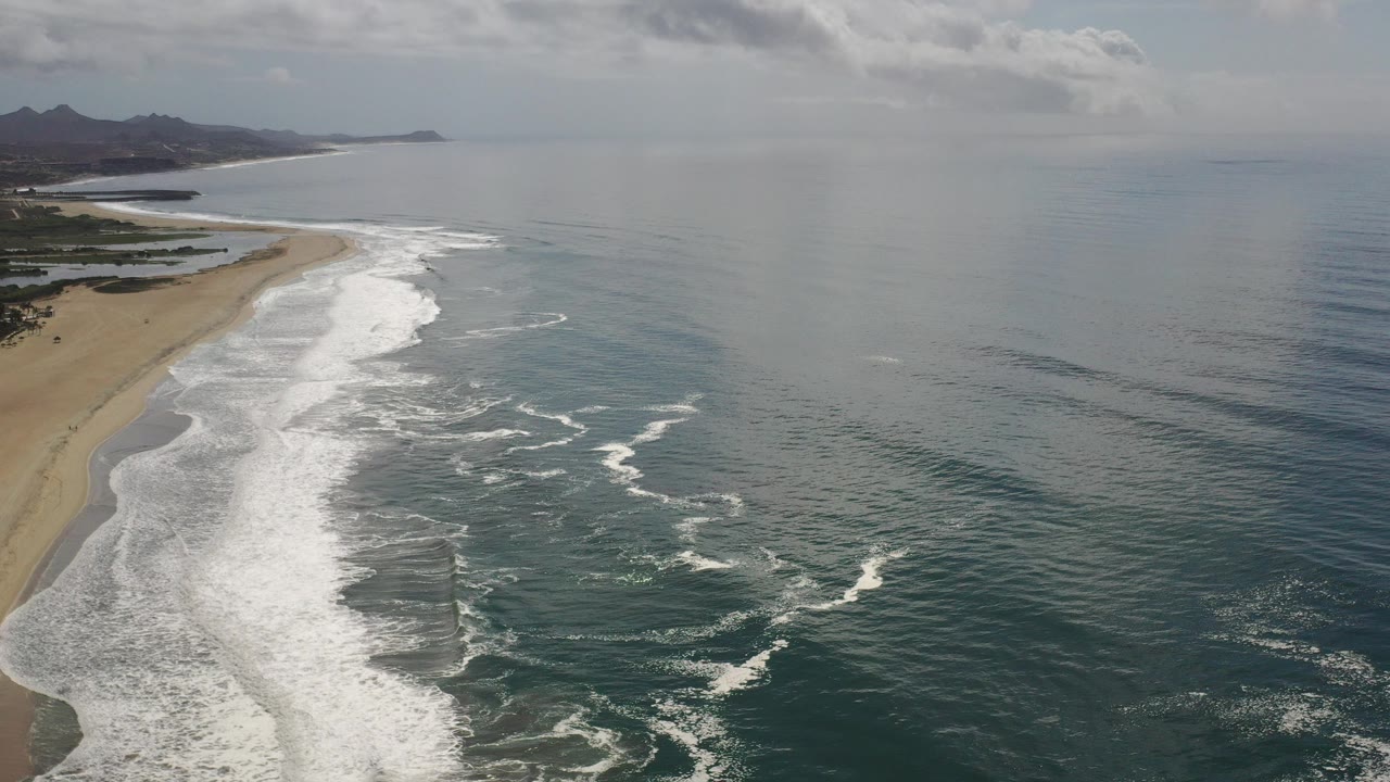Opening aerial view of the Los Cabos coastline with a broad sandy beach on the left, white surf bands along the shore, open ocean to the right, distant mountains, and no visible people.