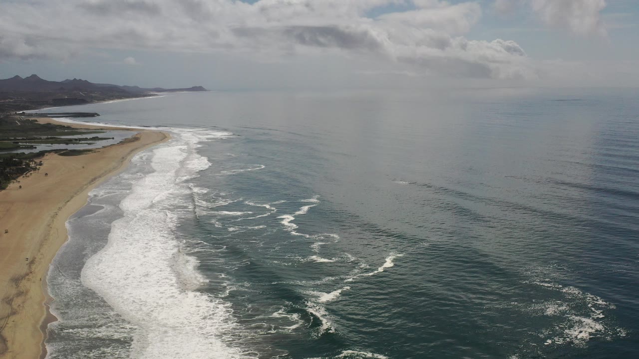 Aerial view over a Los Cabos sandy shoreline with white surf spreading along the left side, blue-green water to the right, distant hills on the horizon, and no visible people.