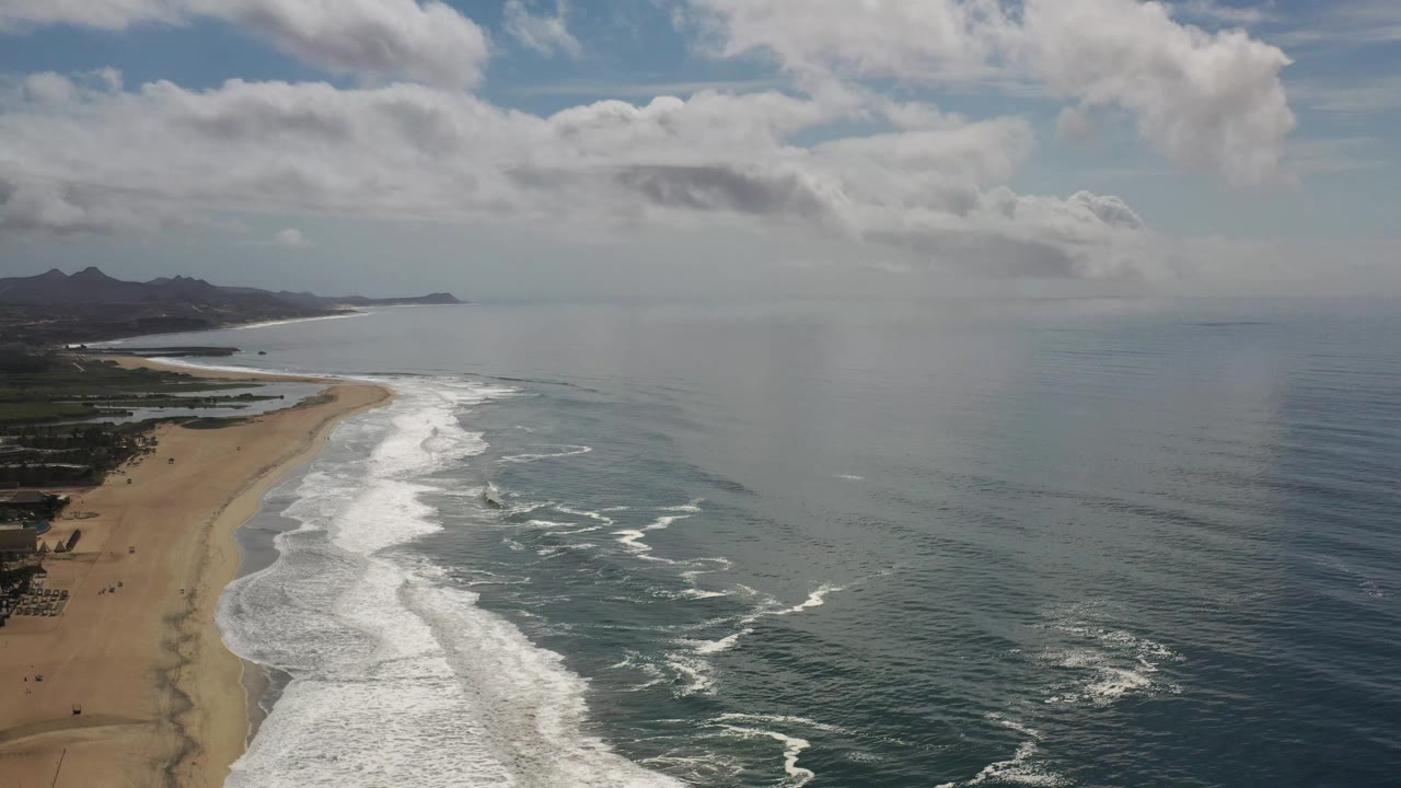 Drone view in Los Cabos showing a long sandy beach on the left, white surf bands in the center, calm blue water to the right, low hills beyond the coast, and no visible people.