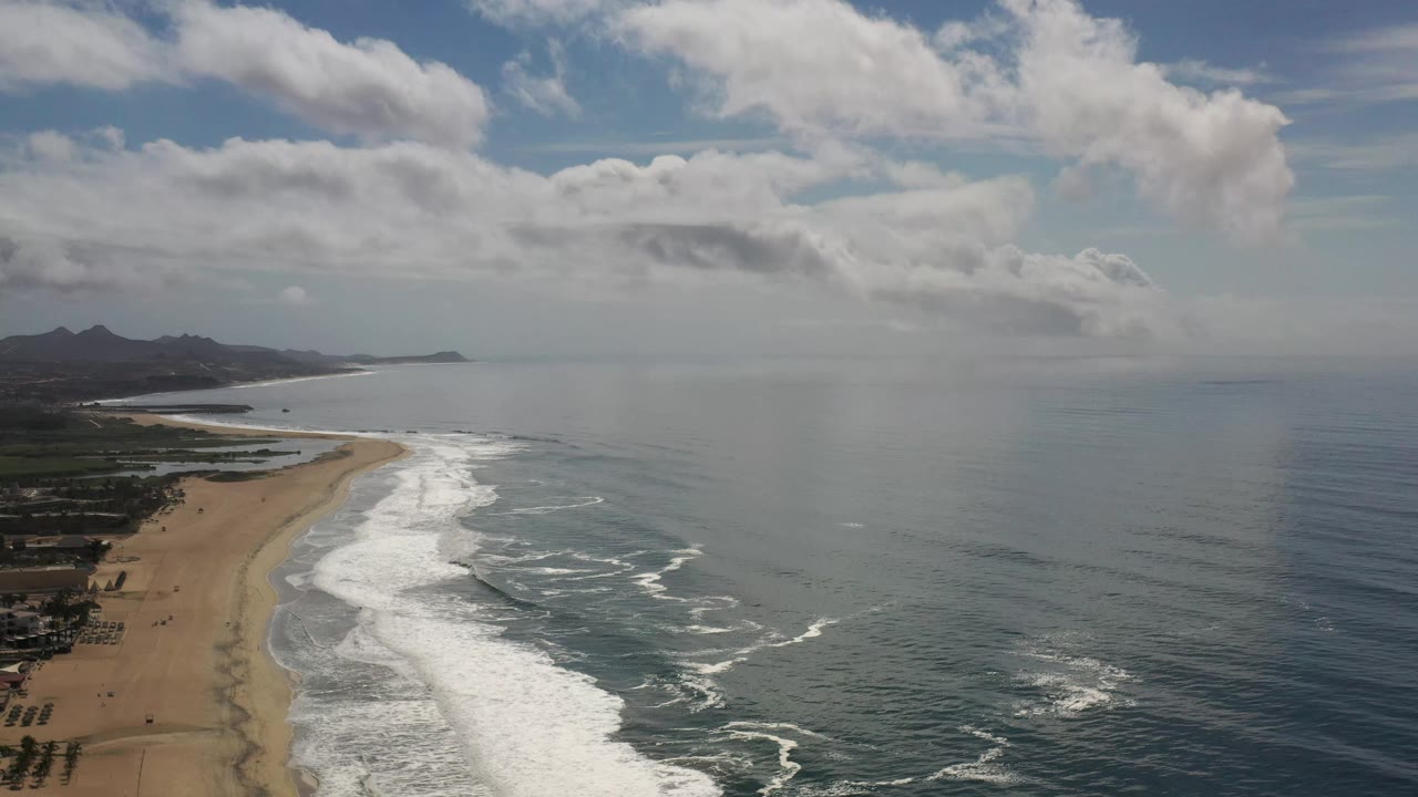 High view of the Los Cabos shoreline as white water spreads farther across the beach edge, with open ocean on the right and no visible people.