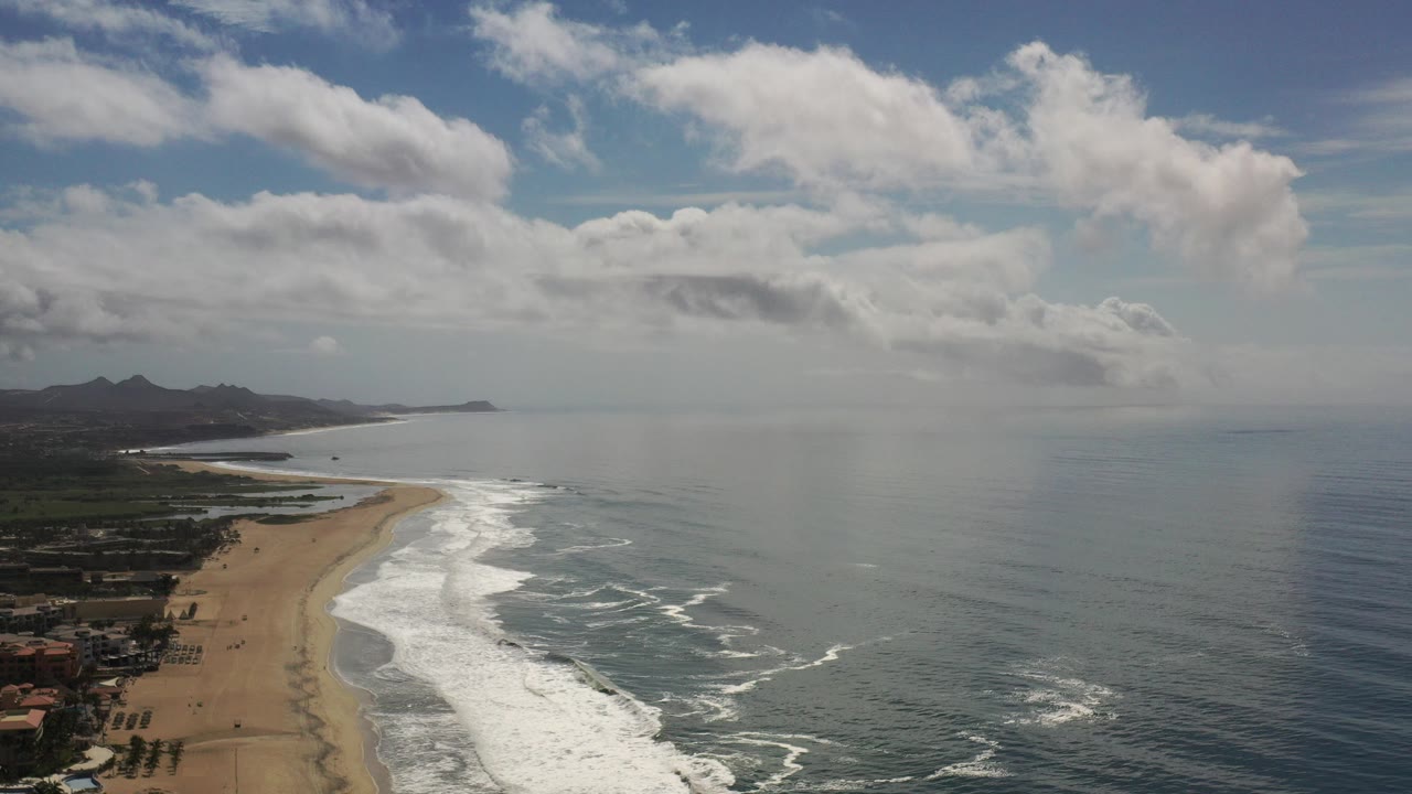 Aerial view over the Los Cabos coast with sandy beach at left, white surf bands, blue ocean to the right, distant mountains, scattered clouds, and no visible people.