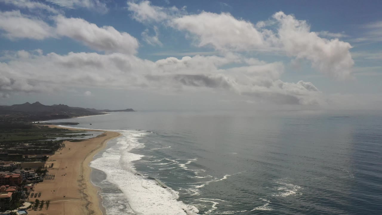 Wide aerial frame of the Los Cabos beach as white waves break along the sand, with coastal buildings low at left, open ocean at right, distant hills, and no visible people.