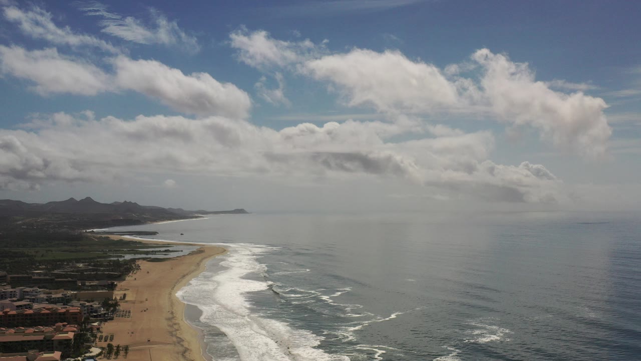 Opening aerial frame over a Los Cabos beach with low buildings at the lower left, white surf along the sand, open ocean to the right, distant hills, scattered clouds, and no visible people.
