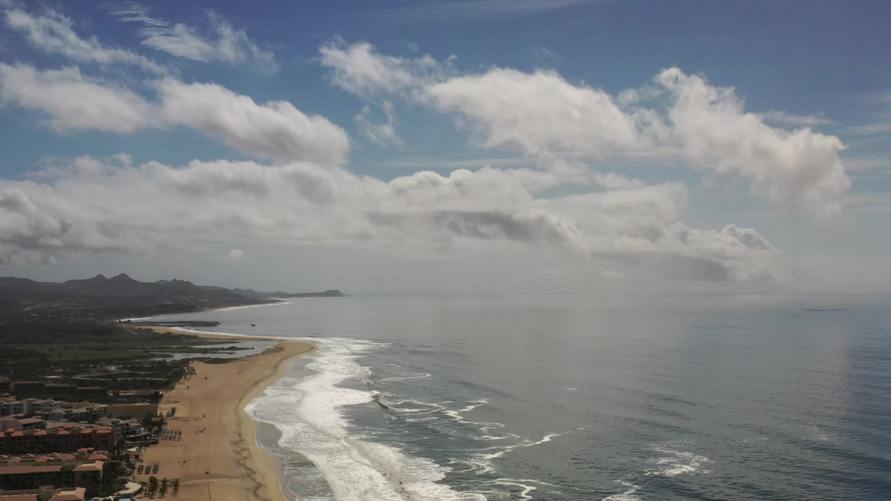 Aerial Los Cabos coastline frame with the beach running from the lower left toward the center, bright surf bands, muted ocean water, distant headland, layered clouds, and no visible people.