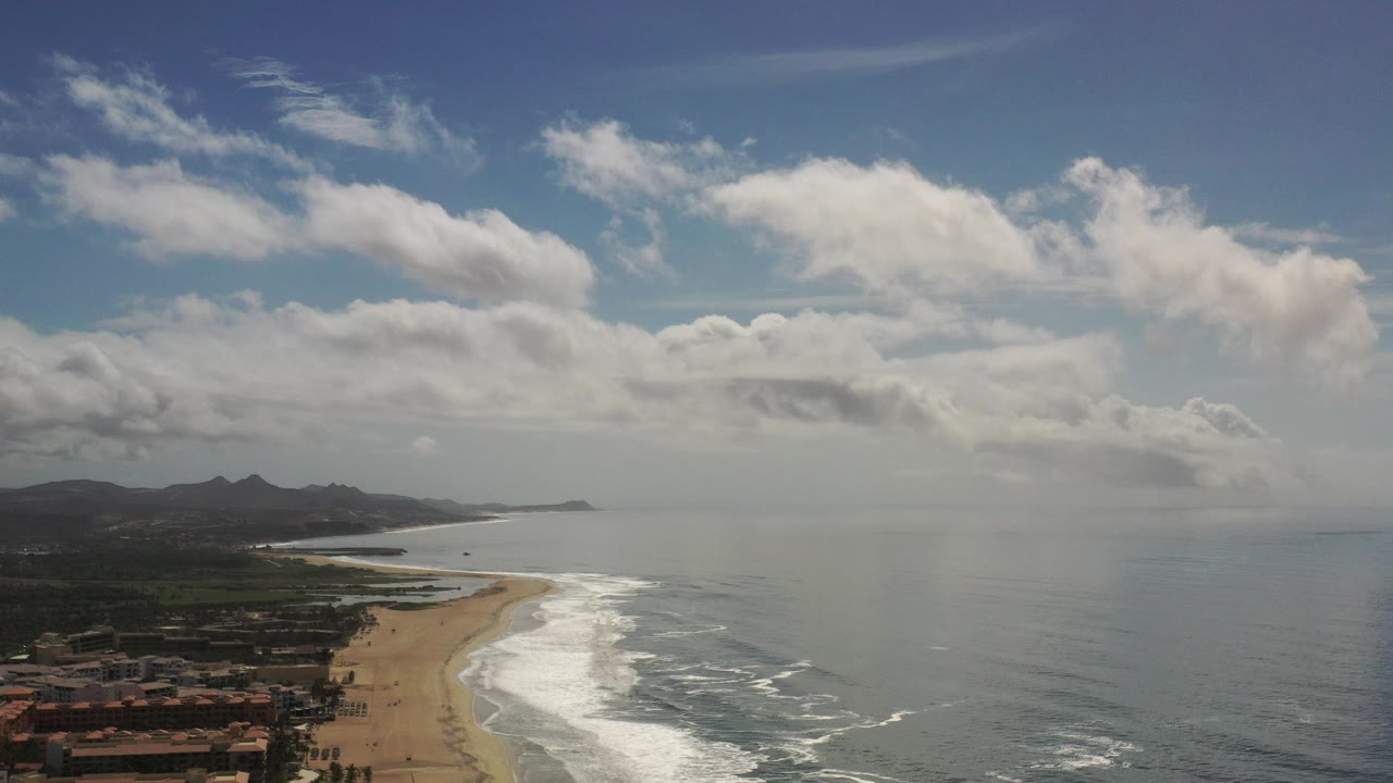 Aerial view over a Los Cabos beach with golden sand at left, white surf along the shore, distant dark mountains, low buildings at the bottom edge, and no visible people.