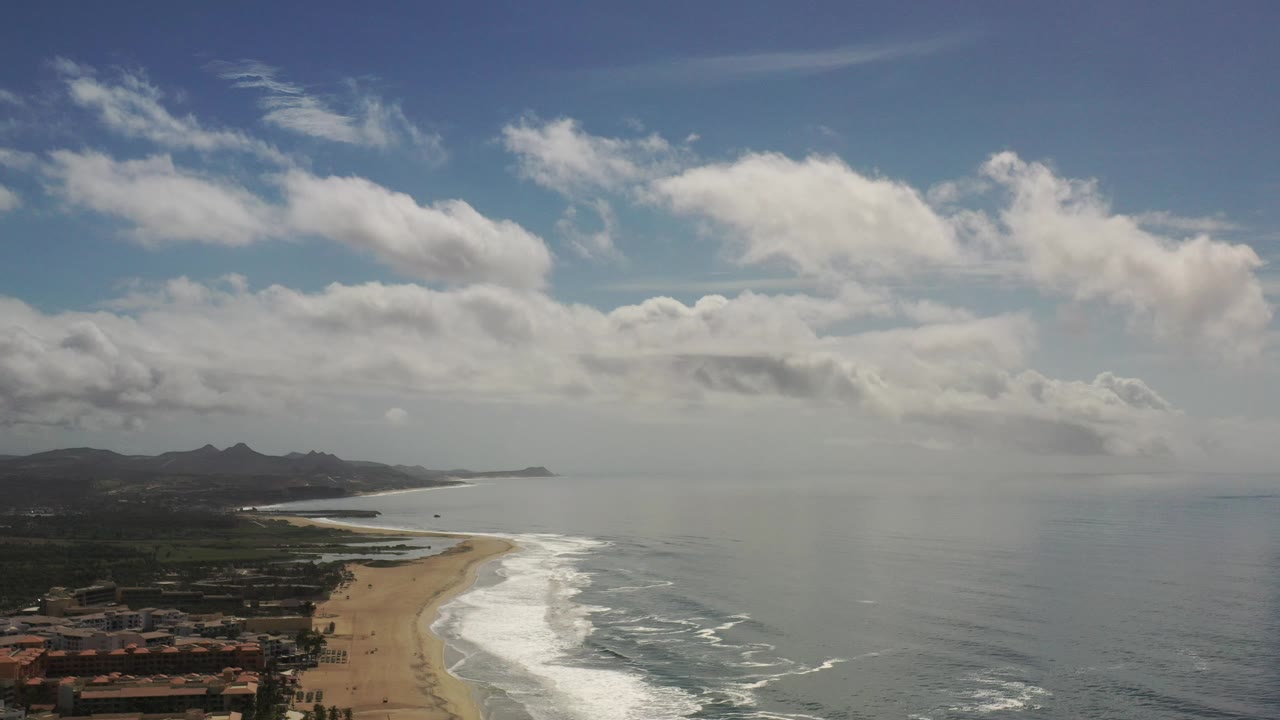 Drone view of sandy coast, ocean, mountain ridge, and scattered clouds.