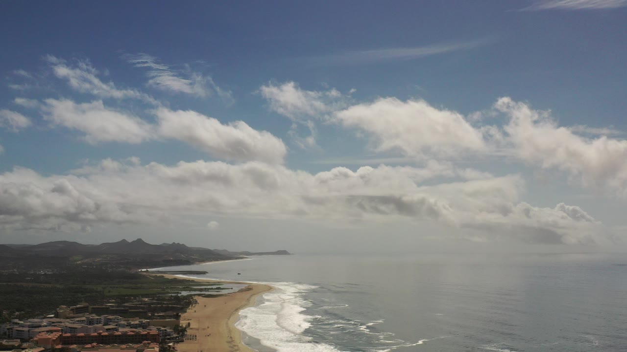 Opening aerial frame over the Los Cabos coast, with a sandy beach at left, white surf lines at center, blue ocean at right, distant mountains, scattered clouds, and no visible people.