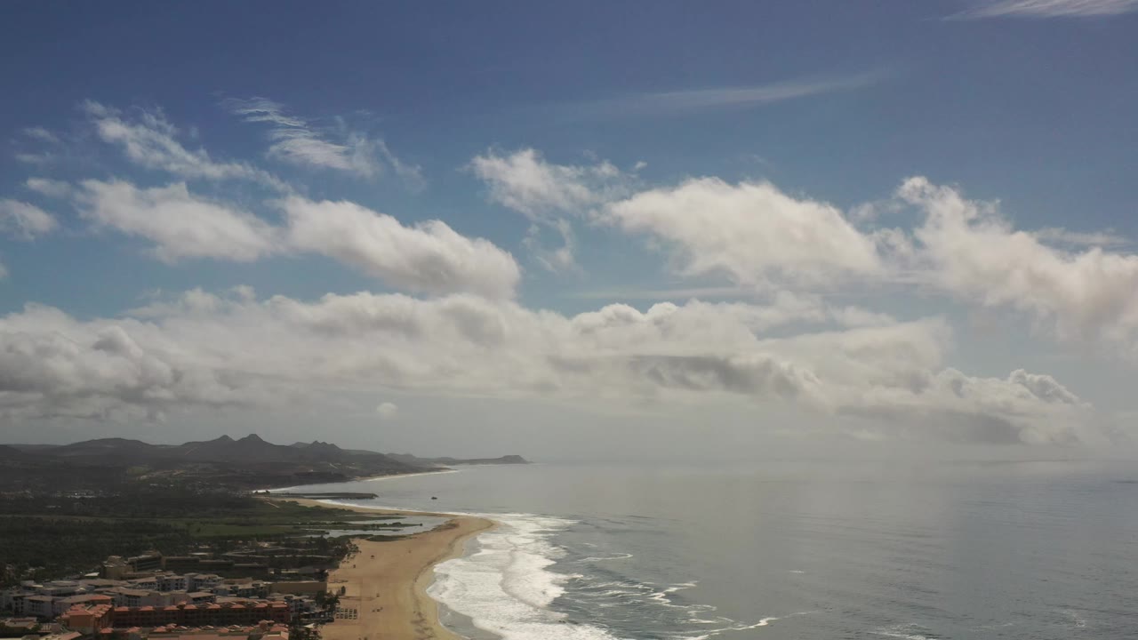 Aerial frame from Los Cabos showing more of the curved beach and breaking waves, with mountains stretching across the left horizon and no visible people.
