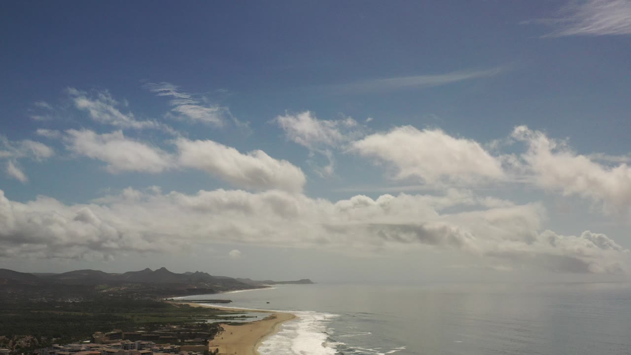 High aerial view of beach, surf, ocean, clouds, and distant hills.