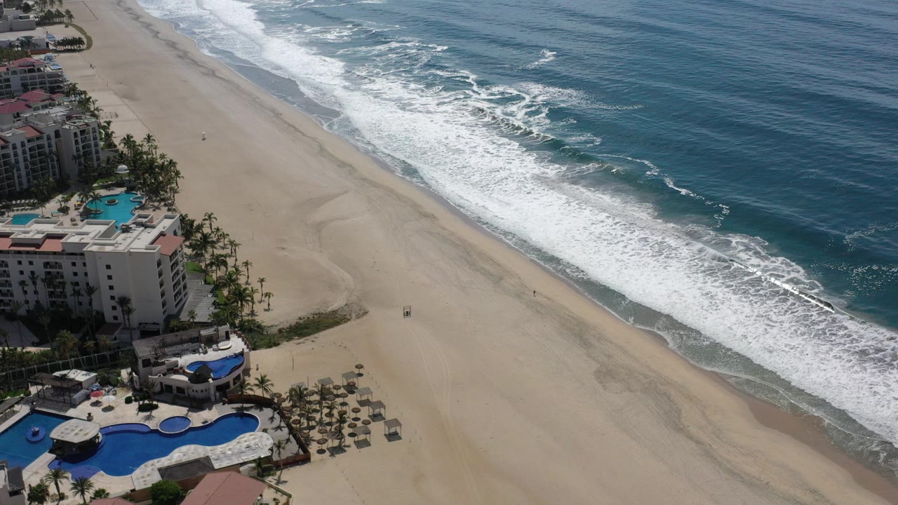 High aerial view over Los Cabos beachfront with blue pools and palm-lined buildings at left, open sand in the middle, breaking waves at right, and no visible people.