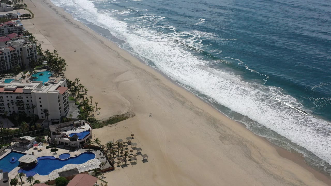 Los Cabos aerial frame showing the shoreline angled across the image, white foam rolling onto tan sand, beachfront buildings and pools on the left, and no visible people.