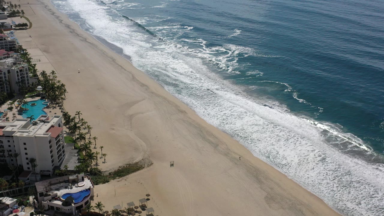 Aerial view of sandy beach, ocean surf, hotel buildings, pools, and palms.