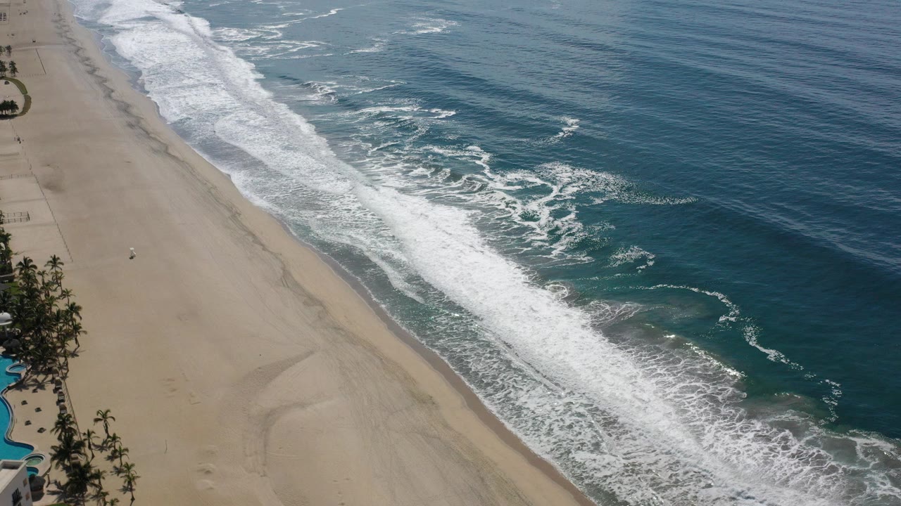 Aerial shoreline with tan sand, white surf, and dark blue ocean.