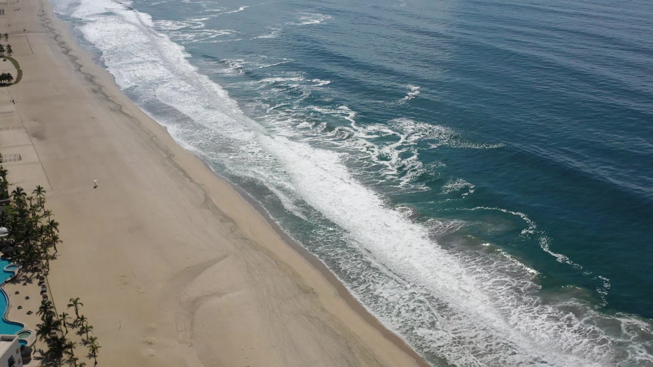Aerial Los Cabos beach frame with a broad white wave band near the sand, turquoise shallows beneath the foam, darker offshore water, and no visible people.