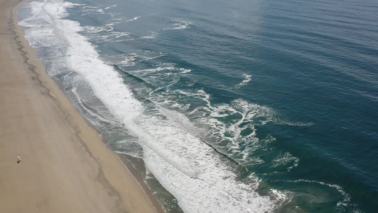 Aerial view over a Los Cabos beach with white surf rolling diagonally along tan sand and dark teal ocean to the right, with no visible people.