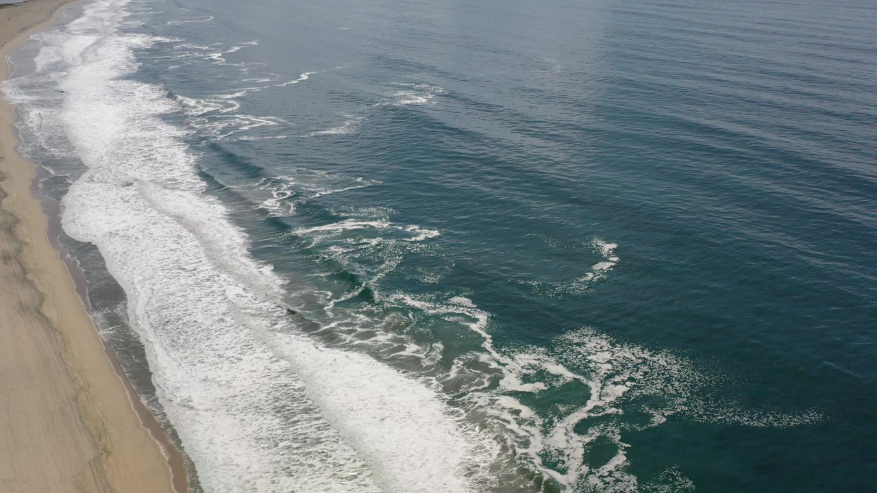 Oblique aerial view over the Los Cabos shoreline with white surf running diagonally beside blue-green water and no visible people.