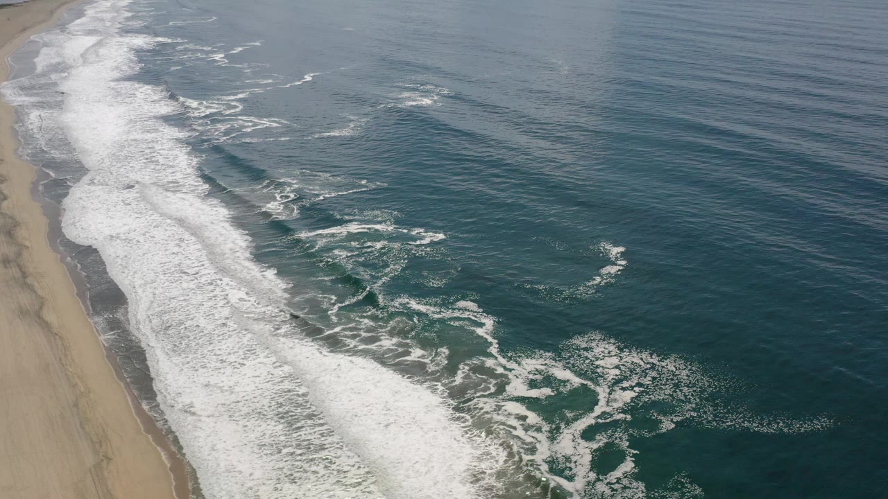Aerial coastal frame in Los Cabos showing a long white breaker near the sand and darker open water to the right, with no visible people.