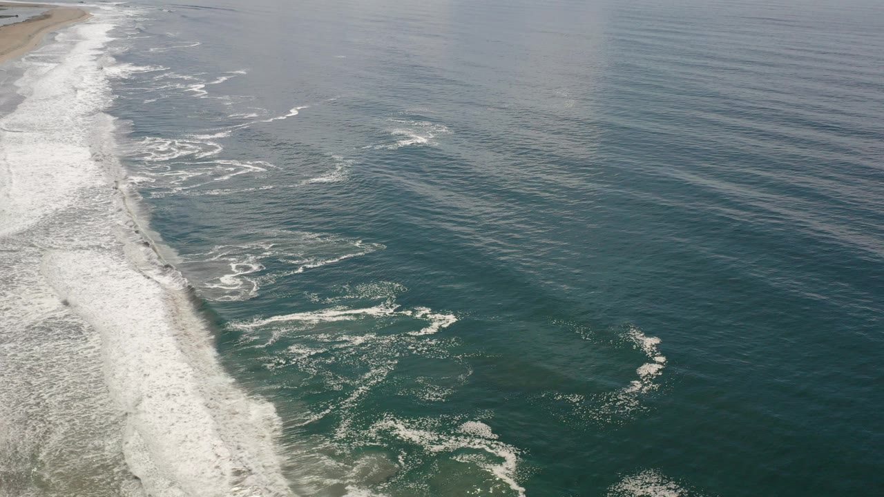 Aerial view over Los Cabos, Mexico, with white surf breaking along the left edge, teal water across the frame, and no visible people.