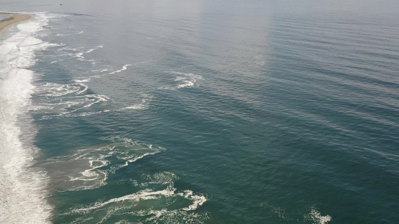 Opening frame of Los Cabos ocean water from above, with white surf breaking along the left edge and no visible people.