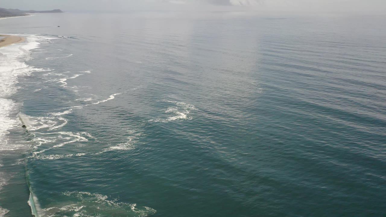 Aerial Los Cabos ocean view with the beach edge sliding along the left side, scattered white foam in the water, a soft gray horizon, and no visible people.