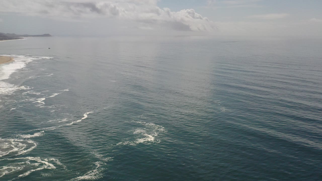 Wide aerial view over the Los Cabos ocean with a sandy beach along the left edge, distant coastline on the horizon, scattered white clouds, and no visible people.