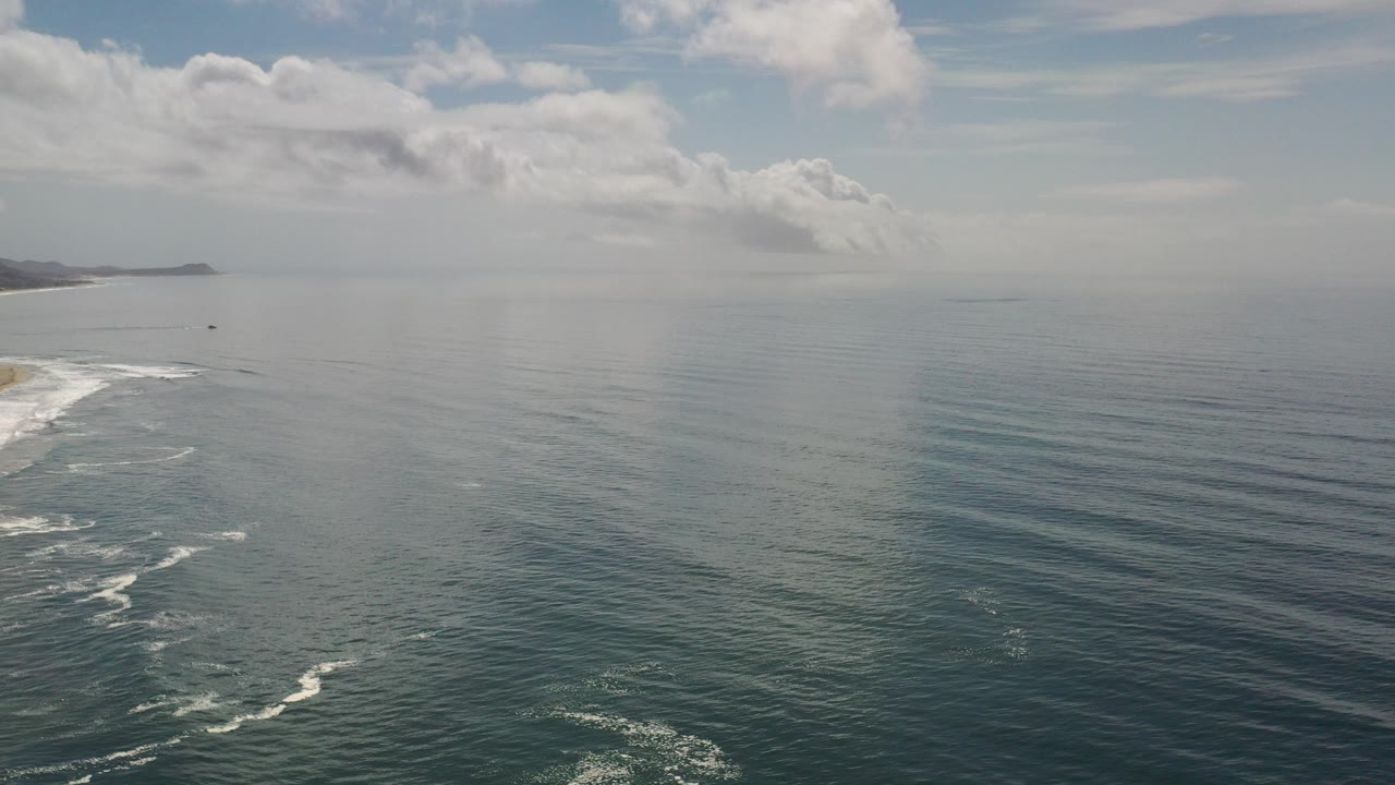 Wide aerial view in Los Cabos, Mexico, with white surf entering from the lower left, open green-blue water, a distant coast on the horizon, scattered clouds, and no visible people.