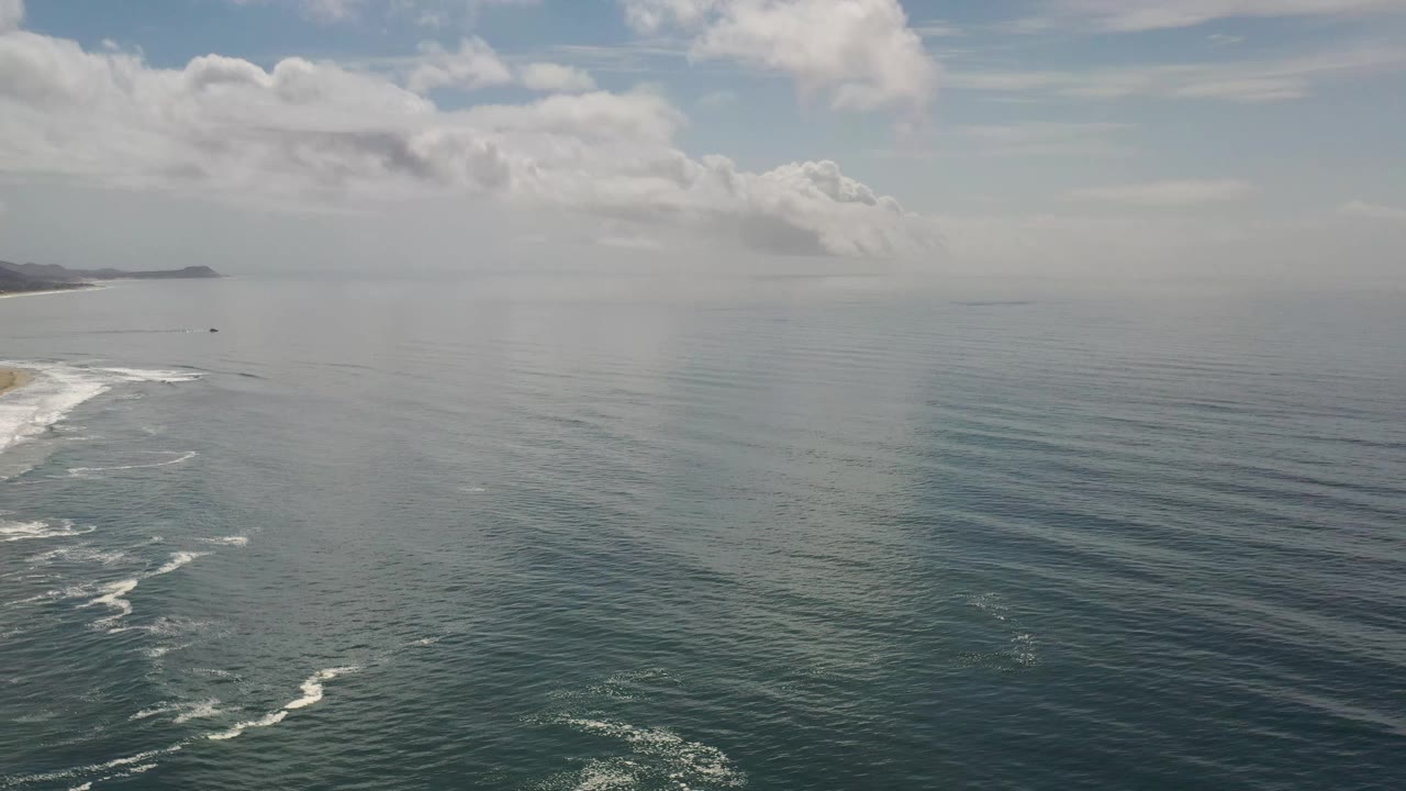 Los Cabos ocean aerial showing more open water across the frame, long parallel swell lines, white surf along the left side, distant land near the horizon, and no visible people.