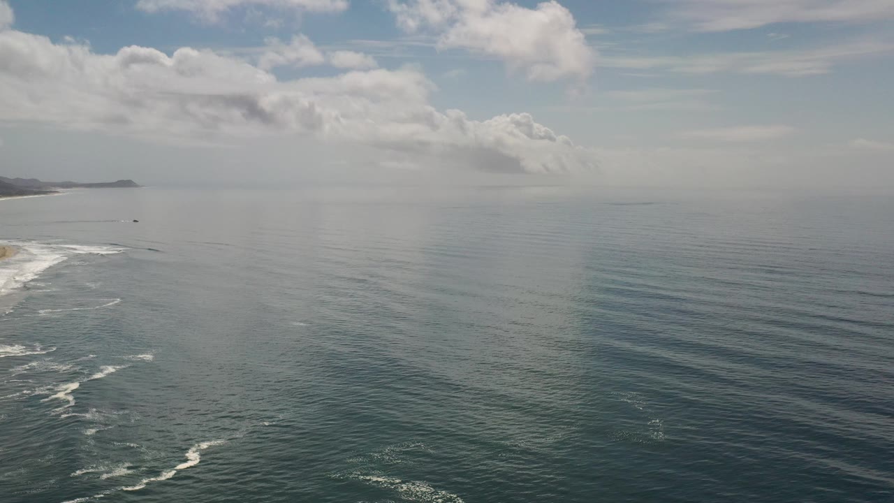 Wide ocean horizon with small surf lines and a clouded sky.