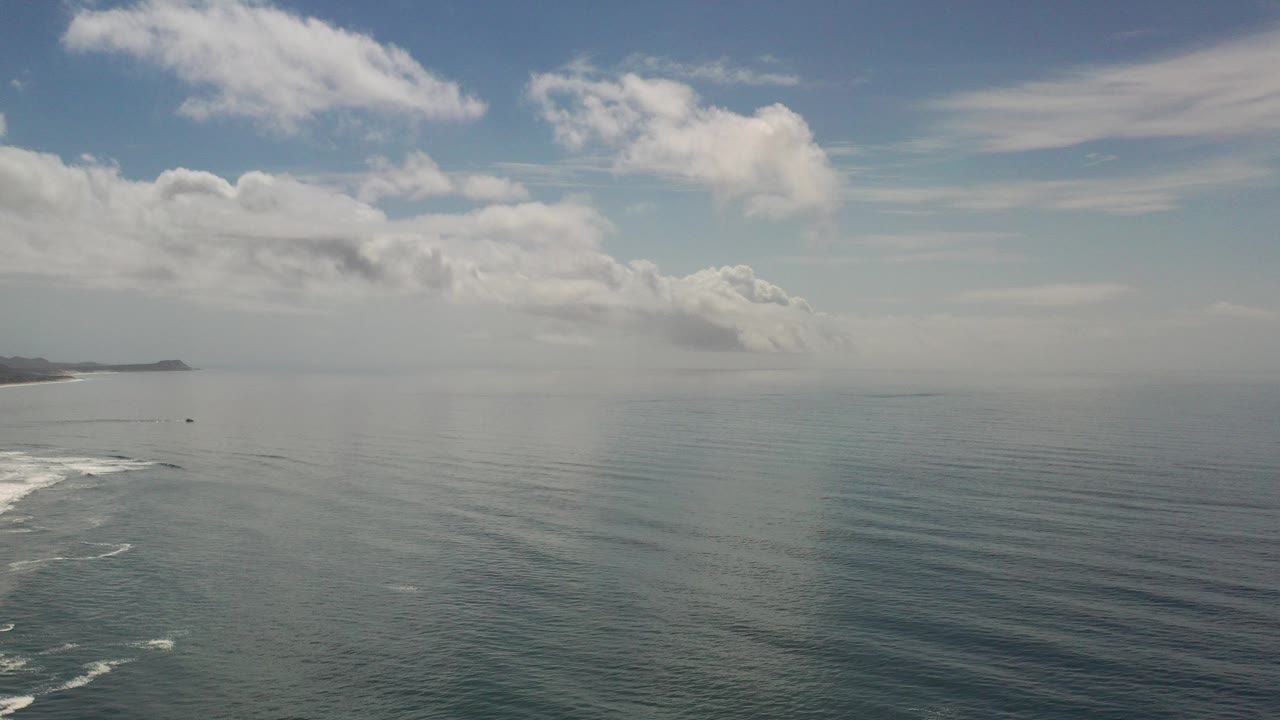 Los Cabos ocean view at the start of the clip, with choppy water, white surf entering from the lower left, distant coastline on the horizon, layered clouds, and no visible people.