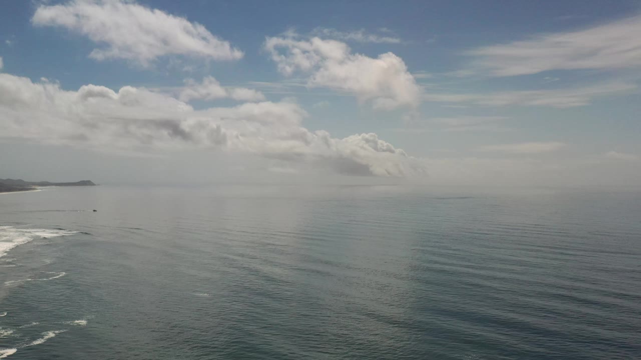 Los Cabos seascape with the ocean surface filling most of the frame, a low distant shore near the horizon, soft cloud bands overhead, and no visible people.