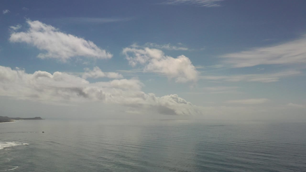 Wide Los Cabos ocean view with scattered white clouds, a distant shoreline on the left, faint surf below, and no visible people.