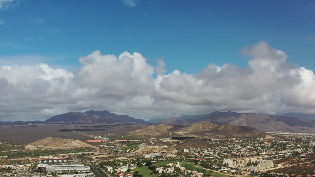 Opening aerial frame over Los Cabos, Mexico, showing low city development in the foreground, dry terrain and mountains beyond, blue sky, large white clouds, and no visible people.