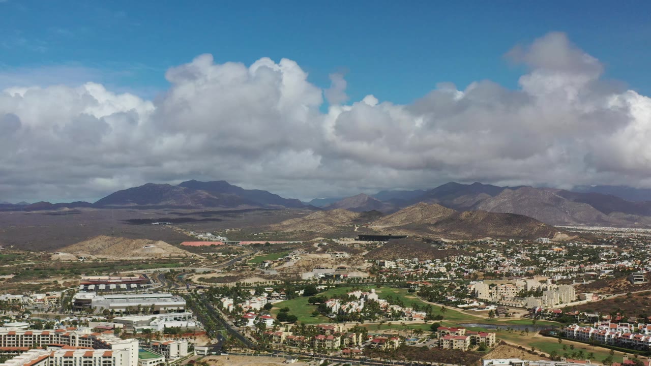 Los Cabos aerial frame with a road corridor, clusters of buildings, maintained grass areas, dry hills, distant mountains, and no visible people.