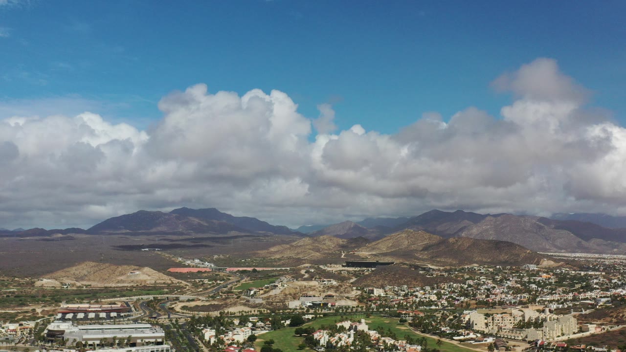 Wide aerial view of buildings below rocky hills and a cloudy blue sky.