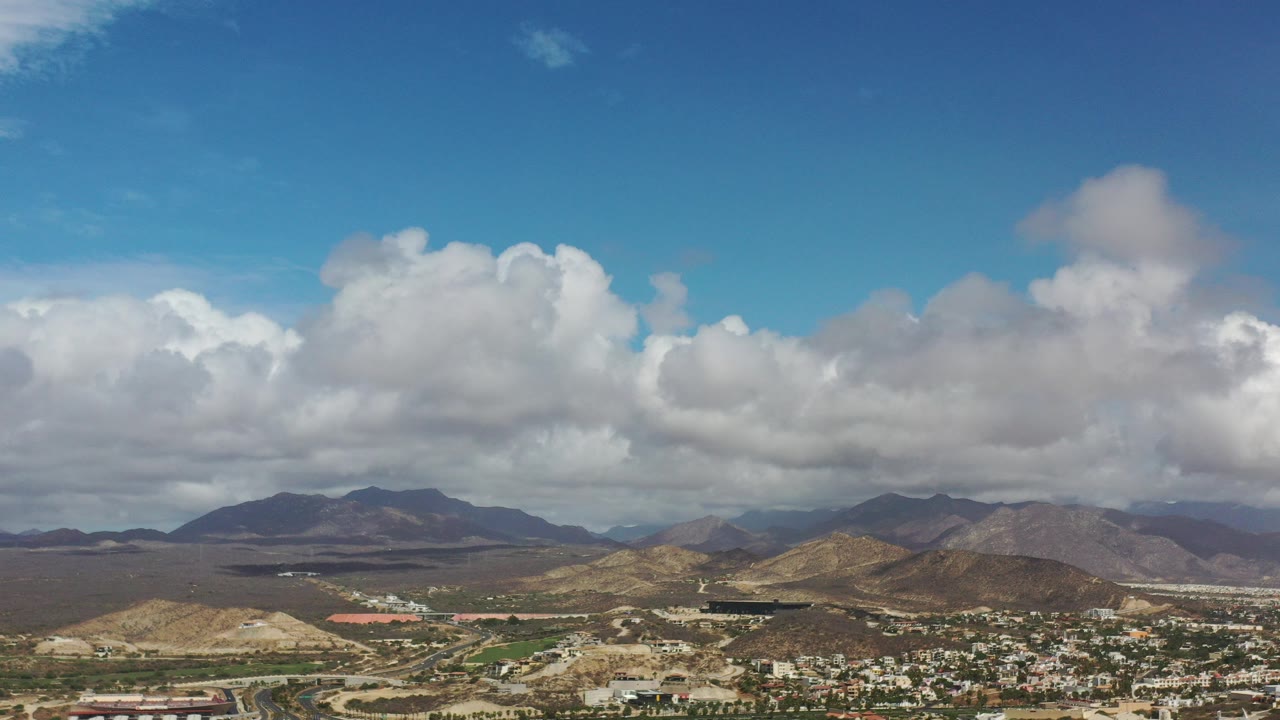 Opening Los Cabos aerial frame with large white clouds over dry hills, distant mountains, roads, and a small town edge, with no visible people.