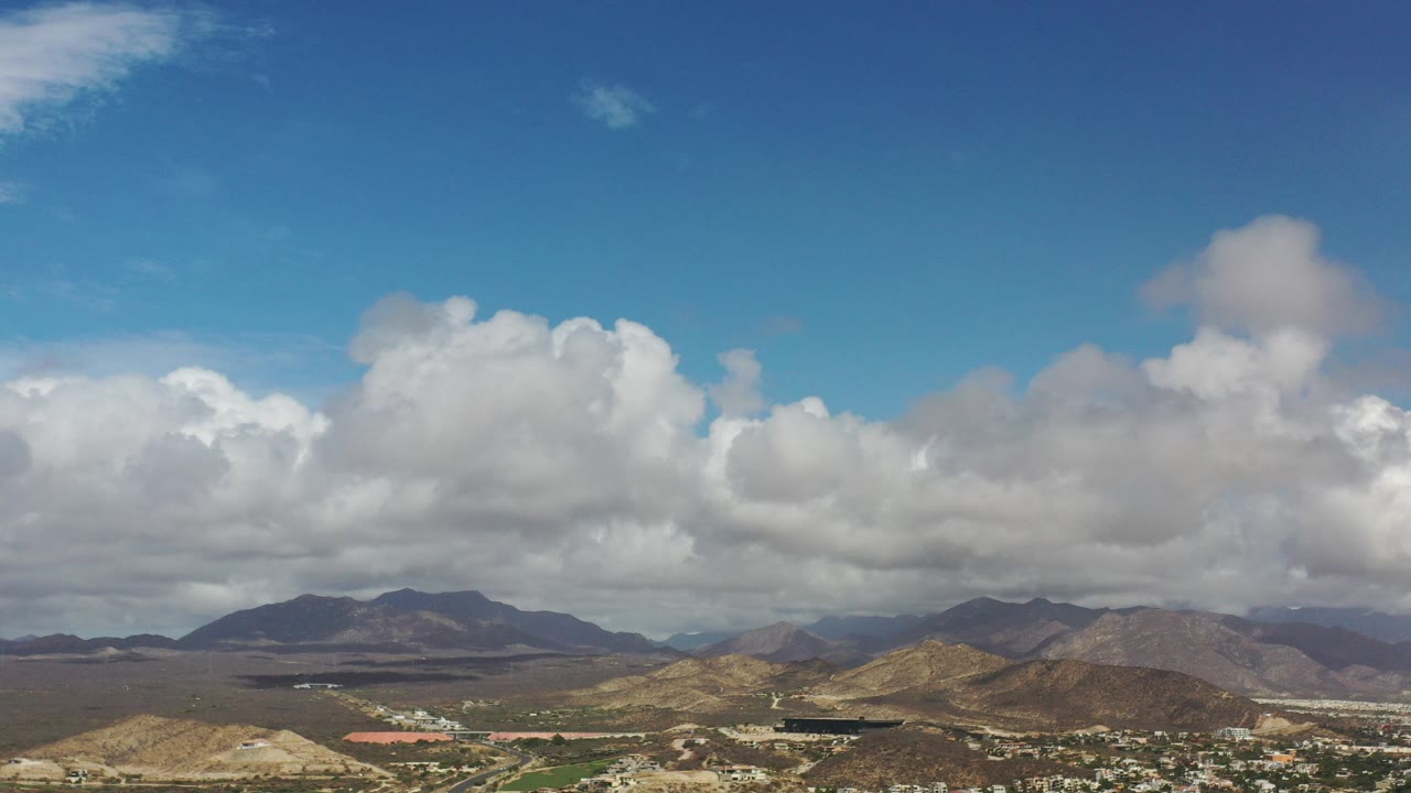 Blue sky and clouds over dry hills, buildings, water, and distant mountains.