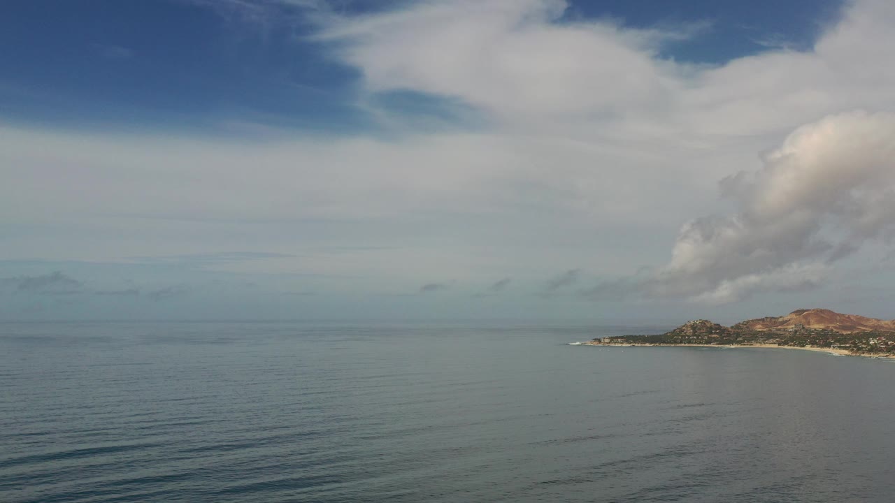 Opening frame of Los Cabos ocean with small dark wave lines in the foreground, a distant island on the right horizon, pale cloud cover above, and no visible people.