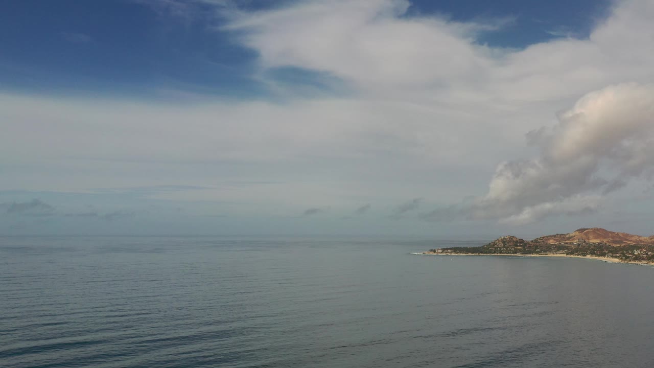 A high view over Los Cabos, Mexico water shows faint surface lines in the foreground, a distant shore to the right, reddish low hills, and no visible people.