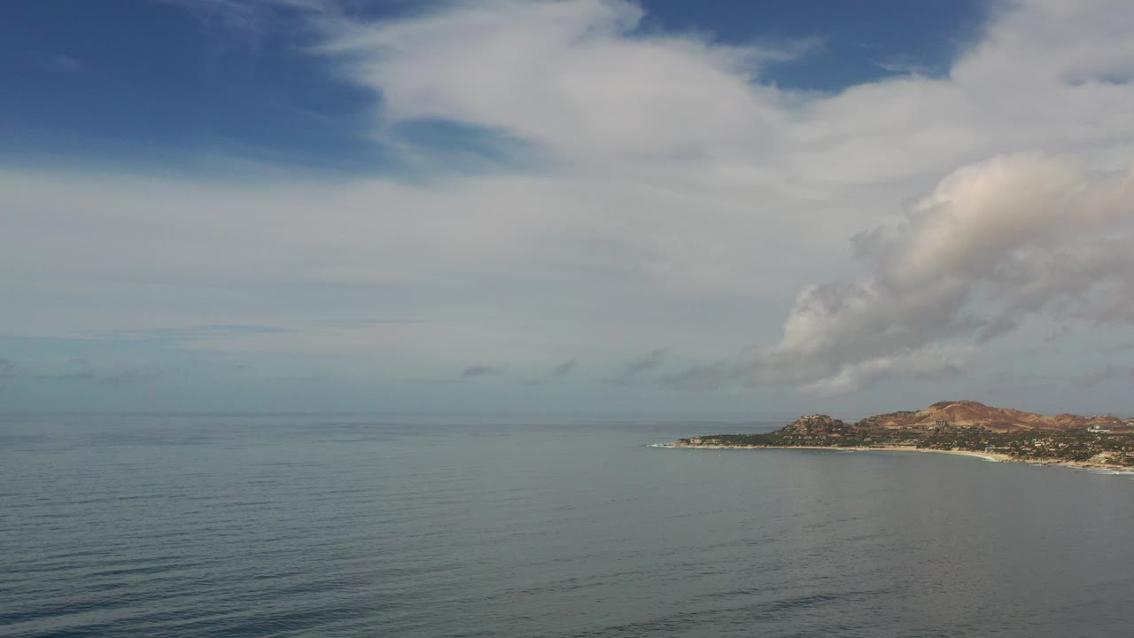 Opening aerial view over bluish water in Los Cabos, Mexico, with a low coastline and reddish hills at right under large clouds, no visible people.