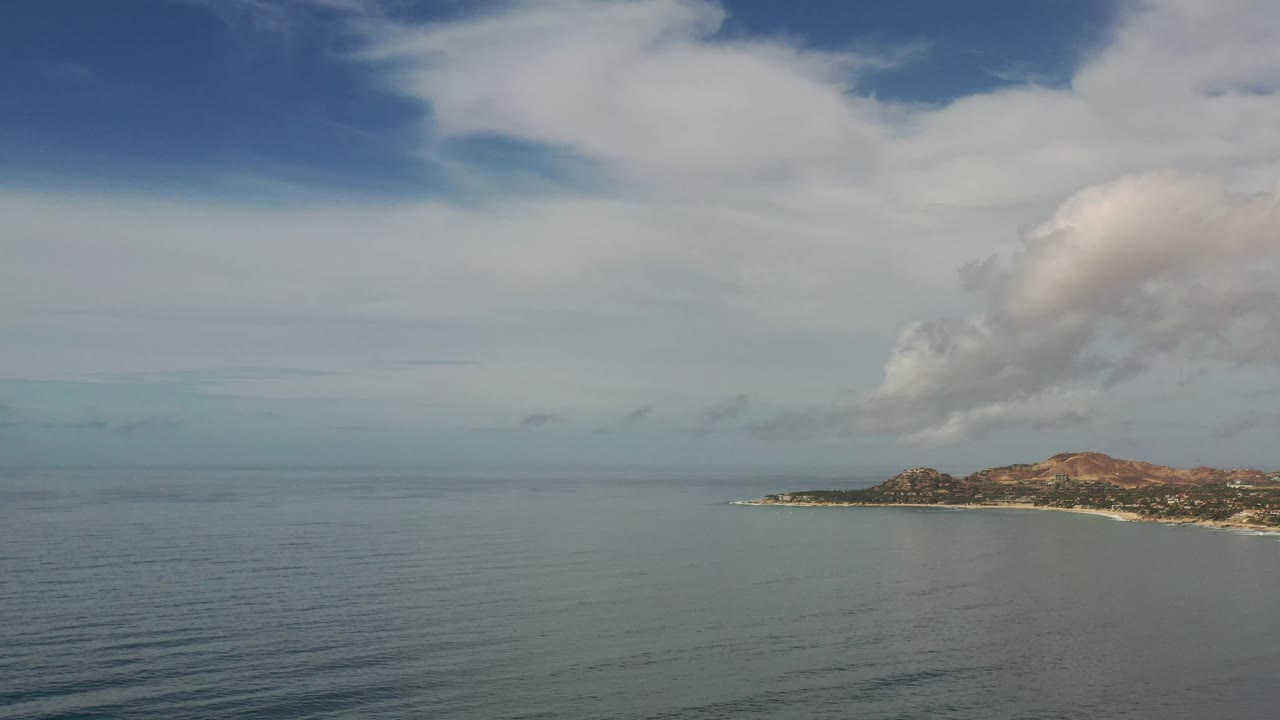 Aerial Los Cabos water view with the shoreline held near the right edge, faint wave lines across the foreground, and no visible people.