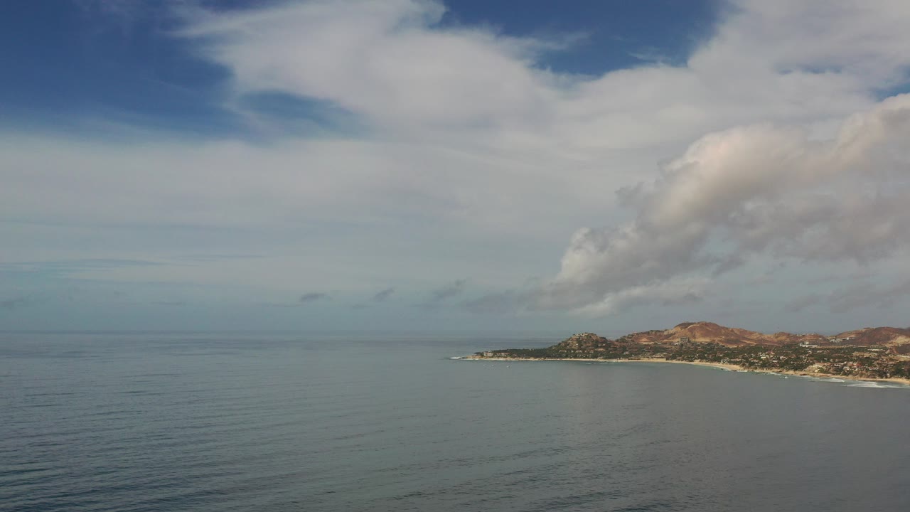 Opening frame of Los Cabos, Mexico with calm sea across the foreground, a low headland and hills on the right, large clouds overhead, and no visible people.