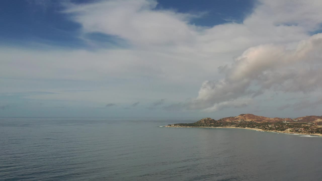 Los Cabos, Mexico coast seen across a broad stretch of blue-green water, with the headland slightly right of center, layered clouds above, and no visible people.