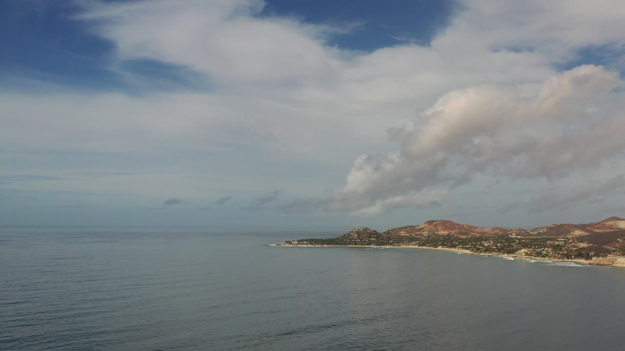 Opening frame of Los Cabos, Mexico from above, with calm blue water in front, low coastline and hills in the distance, broad clouds overhead, and no visible people.